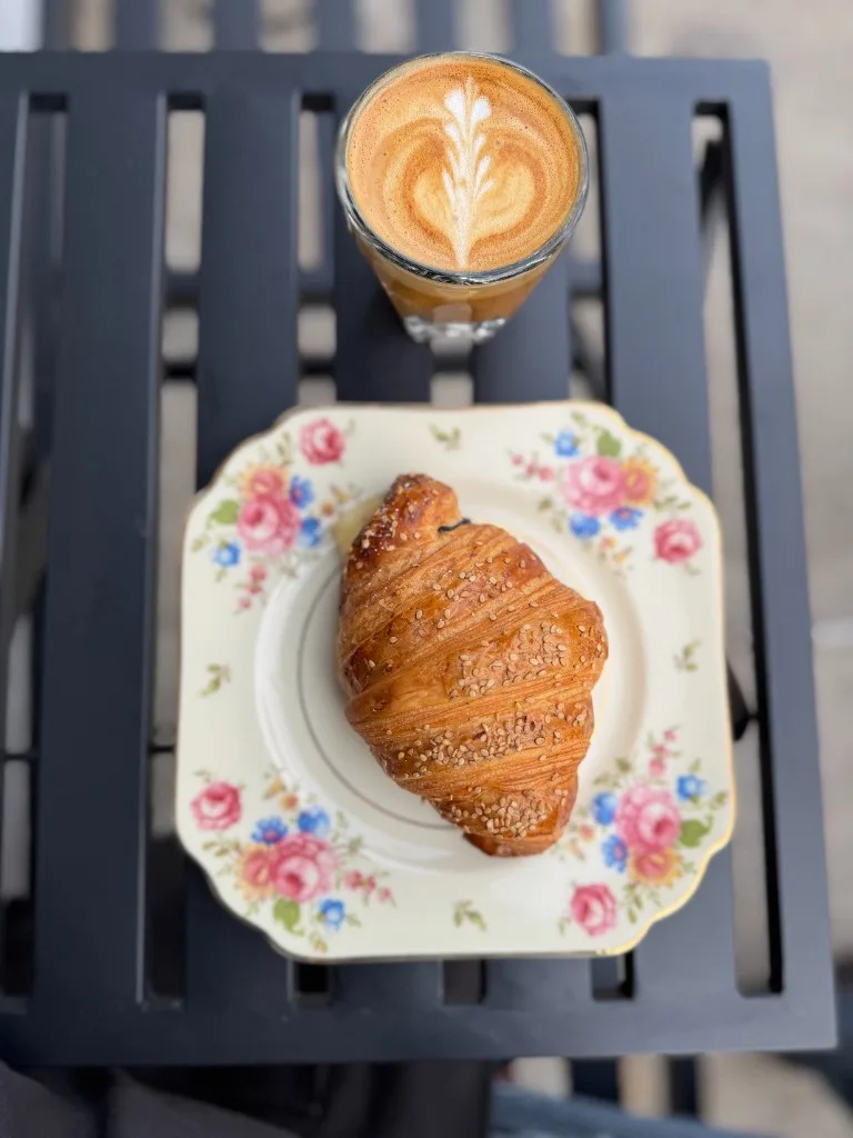 A sesame croissant on a floral plate beside a cortado with latte art