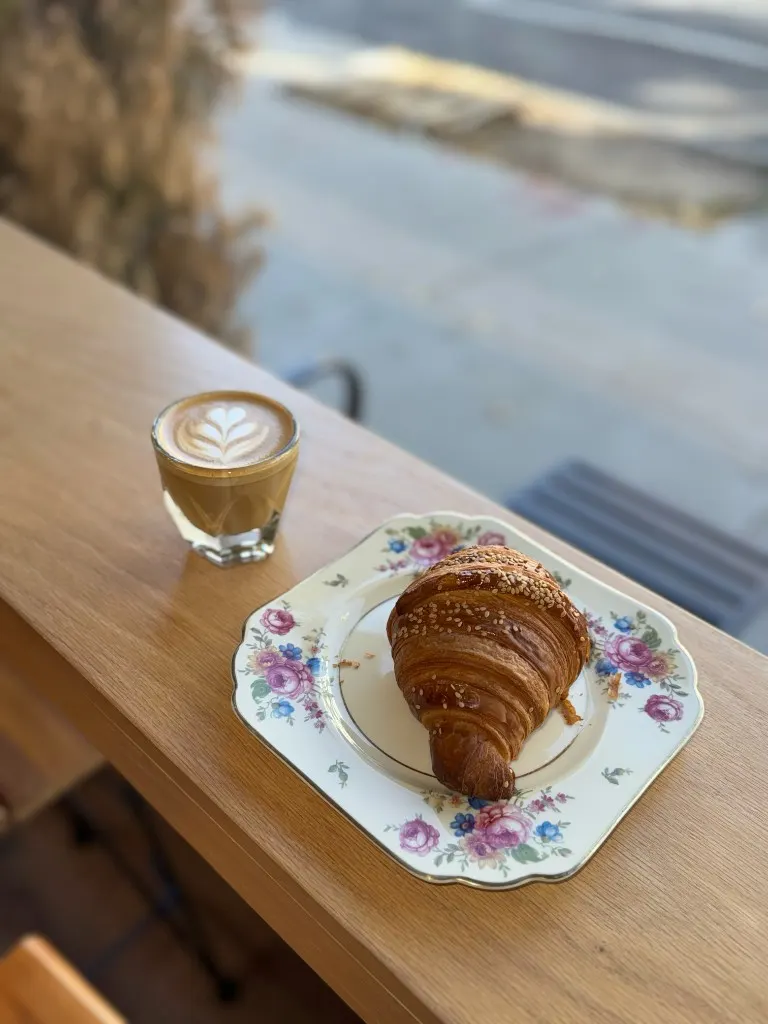 Croissant on a floral plate beside a cortado on a sunlit ledge